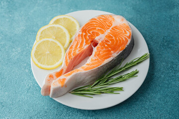 Raw salmon steak with lemon slices and rosemary on white plate over blue background