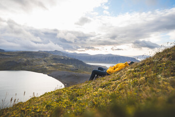 Traveler in a yellow jacket rests on the grassy slopes of The Horn on Snaefellsnes peninsula in Iceland gazing at peaceful lakes cliffs and distant mountains. Chilling on the top of mountain