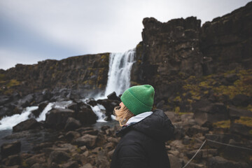 Traveler in a green beanie gazes at Oxararfoss waterfall in Iceland Thingvelli National Park,...