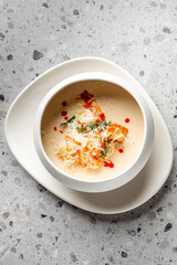 A bowl of creamy soup garnished with shredded cheese, herbs, and a drizzle of red sauce, presented on a white plate atop a textured gray surface. The dish is appetizing and elegantly plated