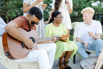 Multiracial group of people, indian man playing guitar and friends eating dinner and drinking wine during party in the picnic