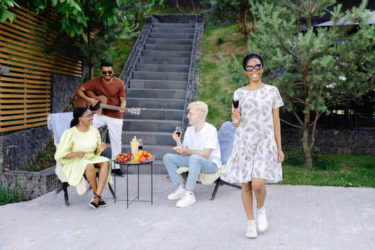 Friends celebrate holiday with wine, fruits and guitar at picnic. Girl with wine goes to the camera and smiles