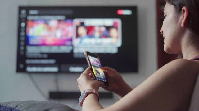 Young woman lounging comfortably on sofa, scrolling smartphone screen with streaming service, large tv displaying matching digital content nearby