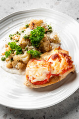 A gourmet dish featuring a baked chicken breast topped with melted cheese and tomato slices, accompanied by a creamy vegetable medley of peas, cauliflower, and potatoes, garnished with fresh parsley