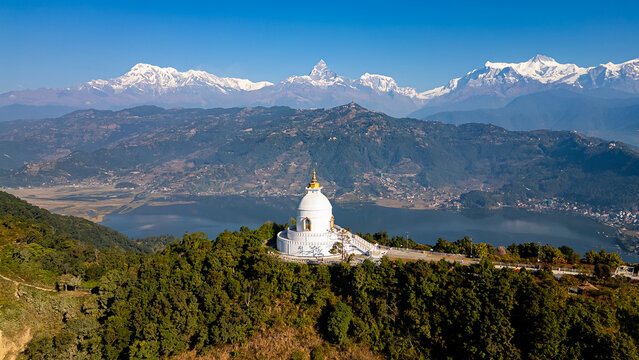 Top view of a white stupa on a green hill, with a calm lake and tall snowy mountains in background.
