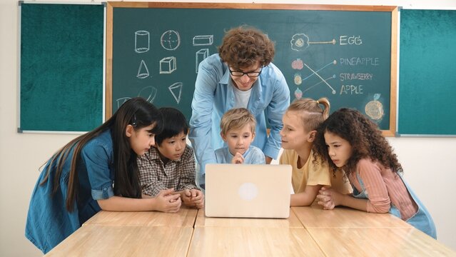 Caucasian teacher and diverse student looking at laptop at classroom. Cute multicultural children learning about coding program or generated program by using software while teacher teaching. Pedagogy.