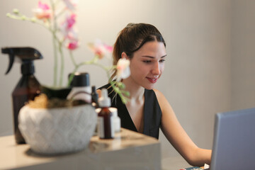 Young woman at a modern reception desk, working on a laptop.  Skincare bottles and fresh flowers suggesting  a spa, beauty salon, wellness clinic, or similar service-oriented space