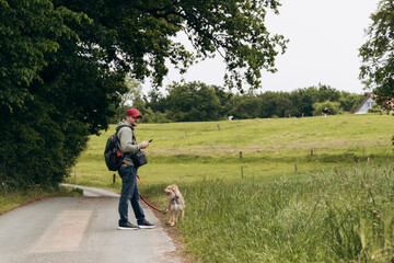Man with backpack and cap stands on rural road, checking phone while walking dog, surrounded by...