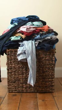 Static medium video of a deeply textured woven wicker basket overflowing with a large, colorful pile of assorted clothes on a rustic wooden plank floor against a cream wall. Time-lapse shows clothes