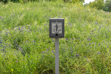Dog poop bag dispenser in a meadow with blooming wildflowers. The label indicates the use of the bags in public spaces.
