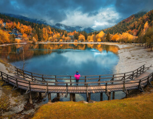Aerial view of woman on wooden bridge, Jasna lake and orange trees at sunset in golden autumn. Kranjska Gora, Slovenia in fall. Top view of bridge, girl, mountains, water, forest, dramatic cloudy sky