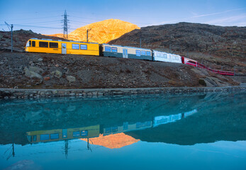Bernina express train passing by Bianco lake with colorful reflections of the train and mountains during sunset in the swiss alps in autumn. Fall in Switzerland. Yellow modern train with red wagons