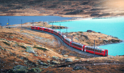 Aerial view of modern red train near azure mountain lake and dramatic cloudy sky at sunset in autumn. Bernina Express, Switzerland. Top view of train in Alps, winding railroad, orange hills in fall