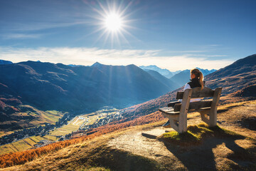 Young woman hiker sitting on the bench on grassy mountain peak enjoying stunning view of valley with high mountain peaks during golden hour sunset in autumn. Hiking in Italy, Livigno. Girl in Alps