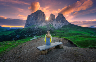 Girl is sitting on the wooden bench on mountain peak in beautiful alpine mountains at sunrise in summer. Landscape with young woman in alps, high rocks, colorful sky with clouds. Dolomites, Italy