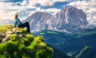 Sitting girl on the top with yellow flowers and green grass in beautiful alpine mountain valley at sunrise in summer. Landscape with relaxing woman in alps, sky with clouds, rocks. Dolomites, Italy