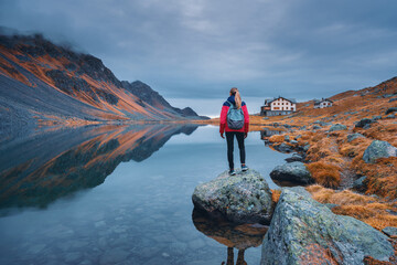 Young woman hiker with backpack on a rock in autumn. Girl and breathtaking view of a mountain alpine lake reflecting the surrounding peaks, orange grass and cloudy dramatic sky. Switzerland in fall