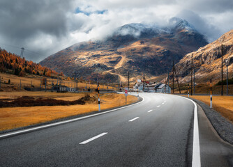 Empty asphalt road winding through alpine mountains in clouds at sunset in autumn. Landscape with majestic Alps, orange trees and grass, dramatic cloudy sky in fall. Road on mountain pass. Switzerland