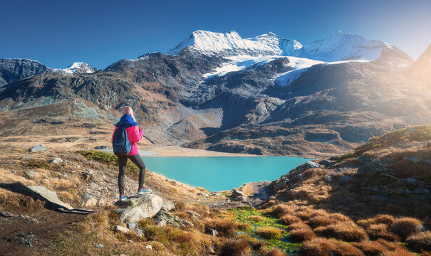 Young woman hiker with backpack standing on a rock admiring a beautiful turquoise alpine lake with snowy mountain and glacier in the background in fall. Girl on mountain trail in Swiss Alps in autumn