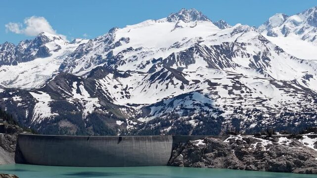 Vue a&eacute;rienne spectaculaire d&rsquo;un barrage en arc au c&oelig;ur des Alpes, entour&eacute; de neige et de montagnes rocheuses. L&rsquo;eau turquoise du lac de retenue contraste avec les parois de b&eacute;ton et la nature sauvage 