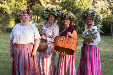 On the evening of the summer solstice, women stand with wreaths on their heads, baskets and flowers in their hands, and colorful clothes. Outdoors, by a campfire.