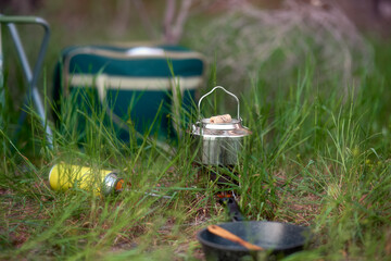 A shiny stainless steel kettle sits on a gas burner in the grass. Summer picnic in the grass.