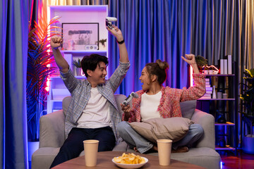 Couple gamer raising fist up holding joysticks be winner together complete level sitting sofa playing video game with front snack and drinks at neon light color living room at modern home. Infobahn.