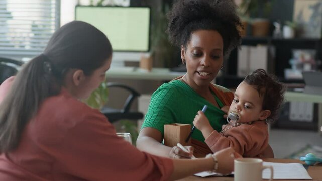 Medium shot of young African American woman with baby son and her female colleague discussing report at office desk