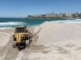 Bulldozer leveling sand on a sunny beach, with a cityscape visible in the background