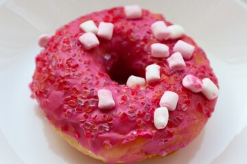 Glazed pink doughnut with marshmallows and colored sprinkles on a white plate, close-up of the dessert on a light background
