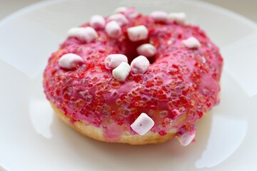 Glazed pink doughnut with marshmallows and colored sprinkles on a white plate, close-up of the dessert on a light background