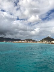 Bright turquoise waters stretch toward a coastal town backed by lush hills under a partly cloudy tropical sky.