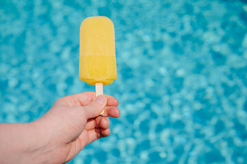 Bright yellow popsicle held above shimmering blue pool water on a warm summer day