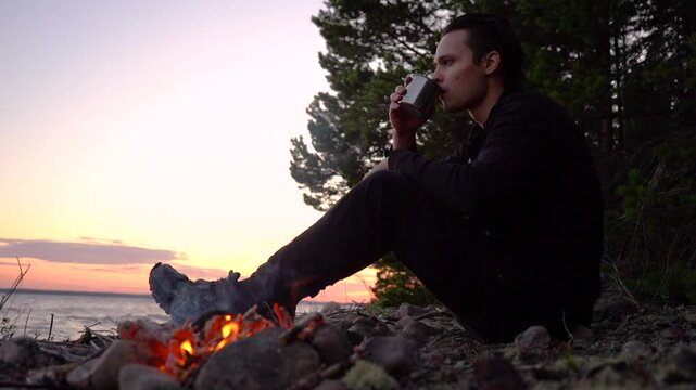  tourist drinks from a mug near a fire on the beach at sunset.