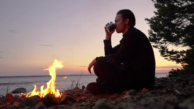  tourist drinks from a mug near a fire on the beach at sunset.