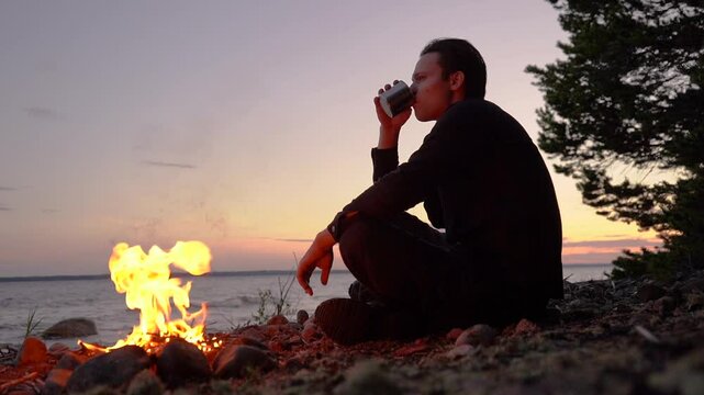  tourist drinks from a mug near a fire on the beach at sunset.