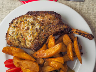 A plate of food with a premium pork chop in the bone, wedges potatoes, and tomatoes. The plate is set on a table with a fork and a knife. Fine tasty dinner with high quality meat.