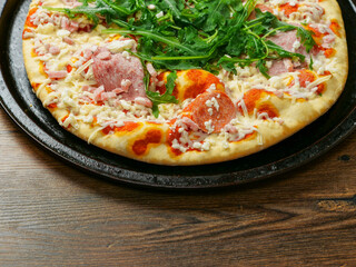 A pizza with ham and spinach on a black tray. The pizza is sitting on a wooden table. Fine Italian cuisine with fresh ingredients ready to be cooked. Fast food meal.