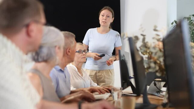 Middle-aged woman teacher making explanation to elderly people sitting at computer table in IT training room