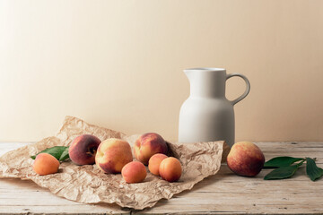 Fresh peaches and apricots with green leaves and ceramic jug on rustic wooden table, minimal beige background. Still life