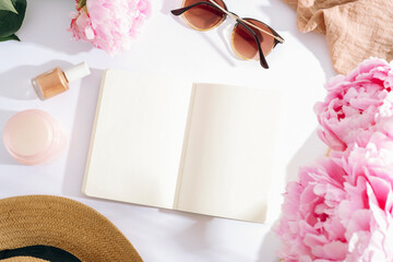 Open notebook with blank pages surrounded by pink peonies, sunglasses, nail polish and straw hat on white background. Top view, flat lay, mockup