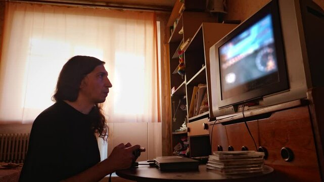 A mid-shot captures a focused and nostalgic mood as a man with long hair plays a video game on an older model television, illuminated by window light, suggesting a classic gaming experience in a home 