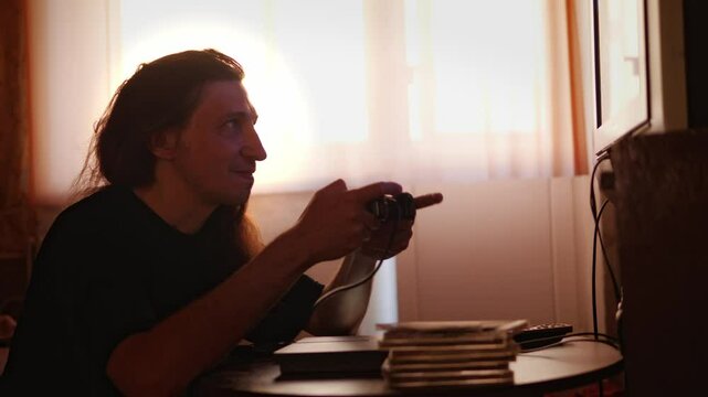 A mid-shot captures an engaged and immersive mood as a man with long hair, backlit by a window, plays a video game with a controller, showcasing a moment of concentration and entertainment at home