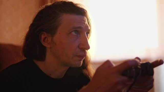 A mid-shot captures an engaged and immersive mood as a man with long hair, backlit by a window, plays a video game with a controller, showcasing a moment of concentration and entertainment at home