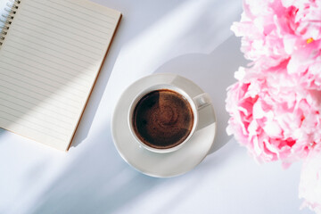A cup of coffee, notepad and peony flowers on white background with shadows. Top view
