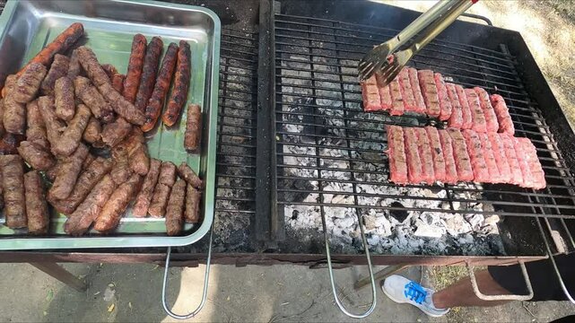 barbecue grill where chevaps (a Balkan dish made from meat) are being fried on coals on a metal grate, a man's hand turning them over