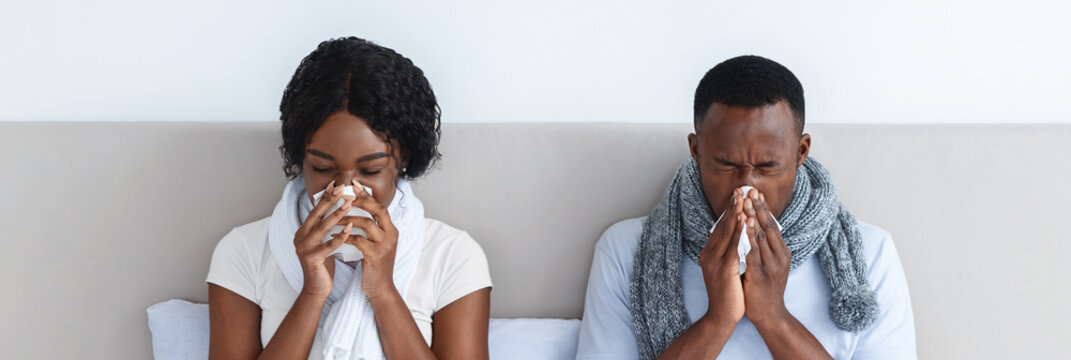 Sick african american man and woman got coronavirus, sneezing their noses, staying in bed during illness. Black couple with scarfs around their necks having flue. Cold and flue concept - Powered by Adobe