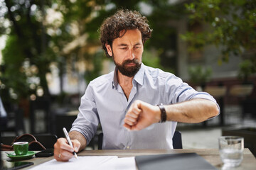A man with curly hair is focused on his work at a café table, writing notes and frequently...