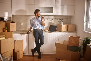 A man stands in a recently moved-in kitchen, sipping coffee while surrounded by boxes,...