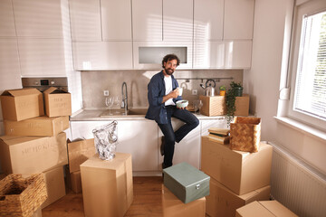 A man sits on a kitchen counter surrounded by packed boxes, holding a cup while contemplating his...